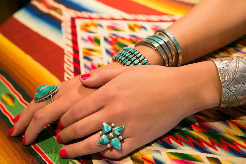 Hands adorned with turquoise rings and silver bangles rest on a colorful woven textile.